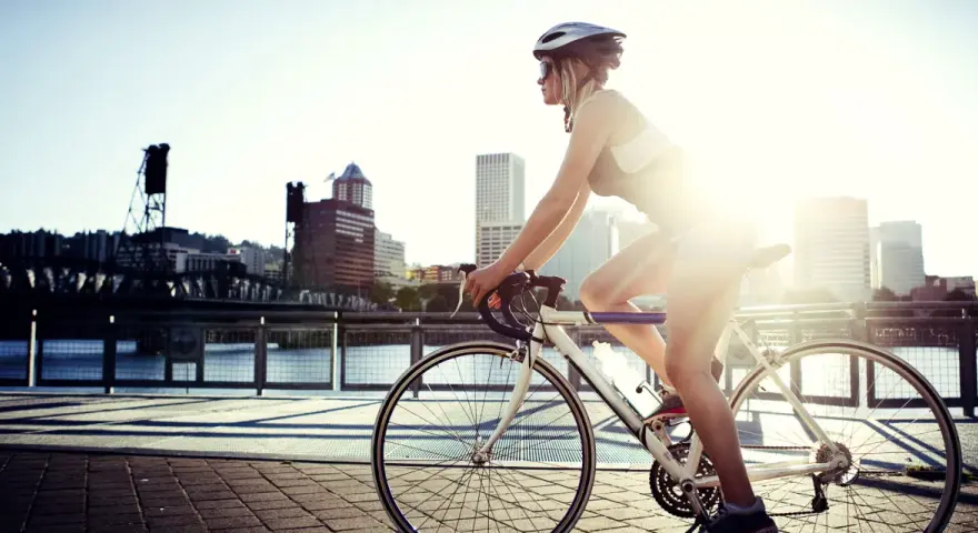 A female cyclist enjoys a sunny ride along a waterfront path with a city skyline in the background.