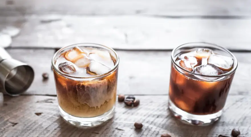 Dwa drinki na drewnianym stole: jeden to kremowy White Russian, drugi to ciemny Black Russian.