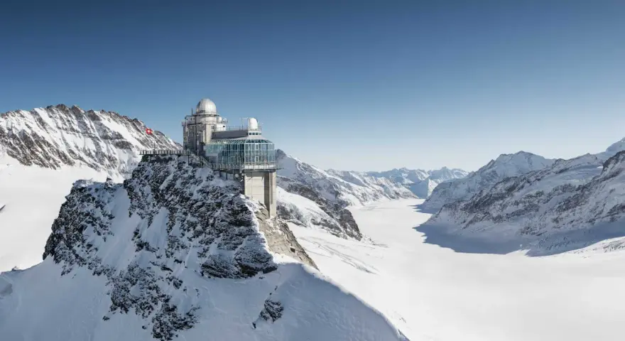 A modern building with glass walls and domes sits atop a snowy mountain, offering breathtaking views of the surrounding peaks and a vast glacier. This is the top of Europe, a place of incredible altitude.