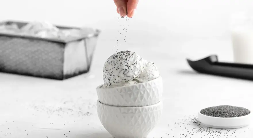 Eine Hand streut Mohn über cremiges Mohn Eis in weißen Schalen. Ein Löffel und eine Schale Mohnsamen liegen daneben.
