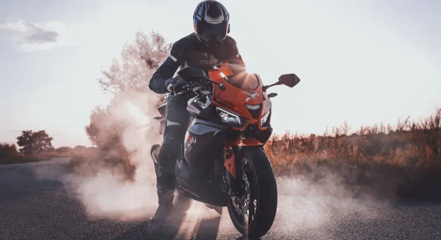 Motocyklista w kasku i kombinezonie na pomarańczowym motocyklu Barton Blade, z opon dymiących na drodze.