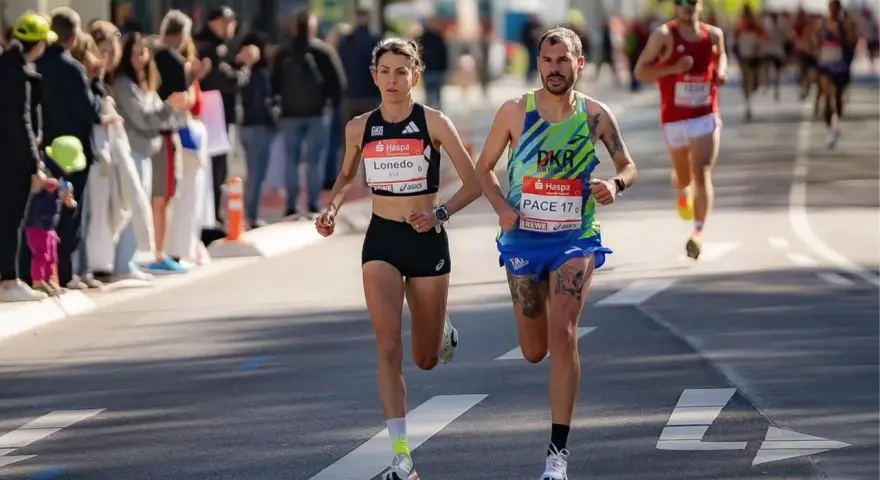 Atleti si preparano a una maratona, correndo fianco a fianco. La folla incita lungo il percorso.