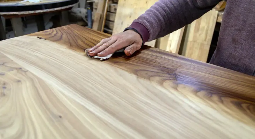 Mano che applica la finitura a cera dopo impregnante su un tavolo di legno grezzo, esaltandone le venature.