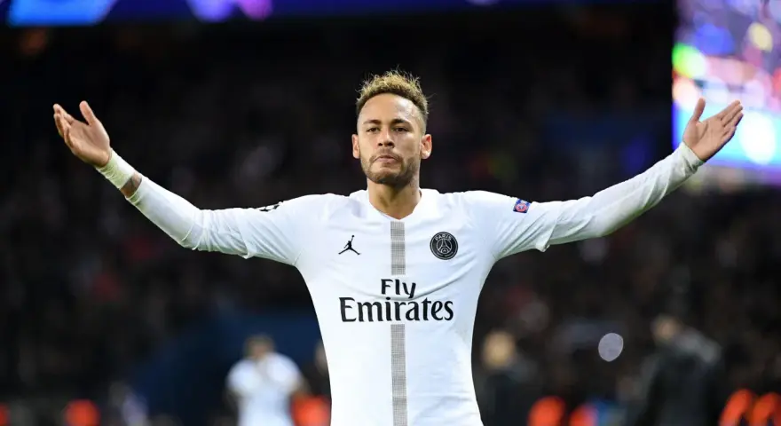 Neymar w białej koszulce PSG, z charakterystyczną neymar fryzurą, unosi ręce w geście radości na tle rozmazanych świateł stadionu.