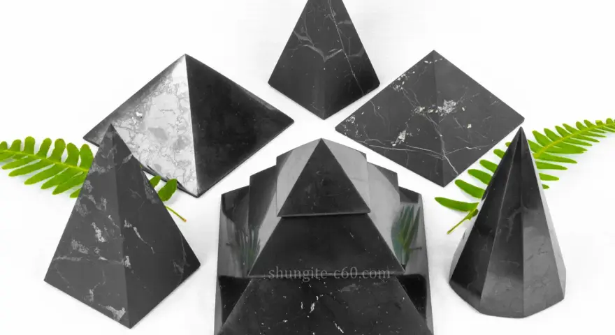 Seis pirámides de piedra shungita, algunas con vetas blancas, dispuestas sobre un fondo blanco con hojas verdes.