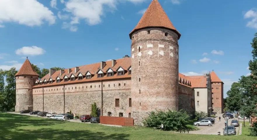 Bytów zamek krzyżacki, masywna budowla z cegły i kamienia z charakterystycznymi wieżami i czerwonymi dachami, otoczona zielenią.
