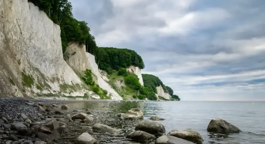 Majestatyczne, białe klify Rugii wznoszą się nad kamienistą plażą i spokojnym morzem. Zielone drzewa porastają szczyty, a chmury tworzą dramatyczne tło.