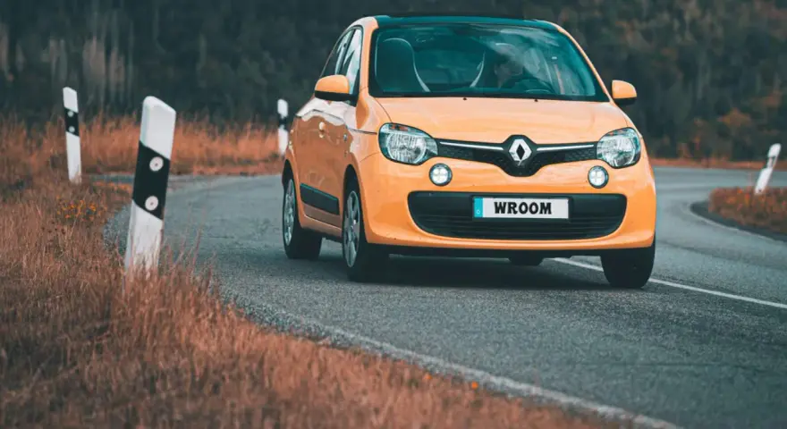 Une Twingo orange roule sur une route sinueuse bordée d'herbes sèches.