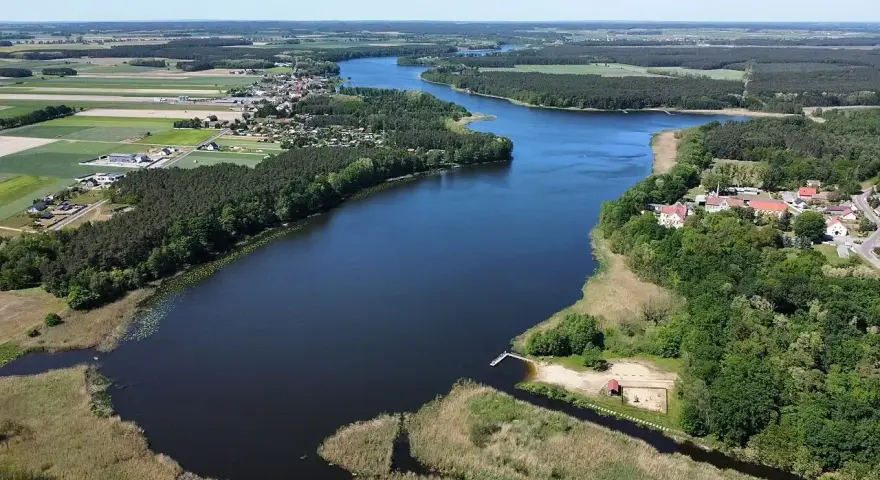 Malownicze jezioro Wojnowskie otoczone lasami i polami. W oddali widać zabudowania wsi, a na brzegu plażę z pomostem.