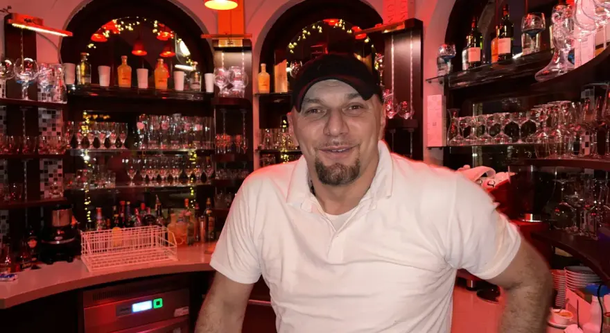 Mann mit Bart und Kappe hinter der Bar, umgeben von Gläsern und Flaschen. Er lächelt freundlich, bereit für die nächste Bestellung in "la gondola due".