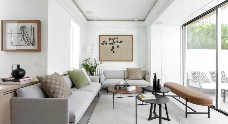 Salón moderno con sofás grises, mesas bajas negras y marrones, y un banco de cuero. Grandes ventanales conectan el interior con la terraza, creando un espacio fluido para unir terraza y salón.