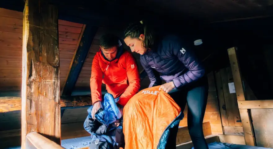 Préparation pour une nuit en refuge. Un homme en veste rouge et une femme en veste violette déballent des sacs de couchage dans une cabane en bois.