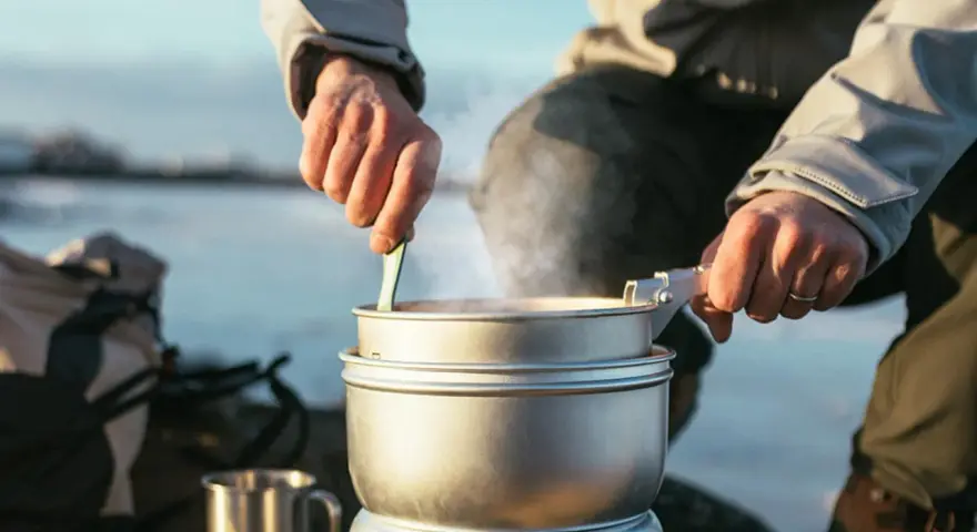 En person lagar mat utomhus, ångande kastruller och en mugg i metall antyder en lyxig matupplevelse i naturen.