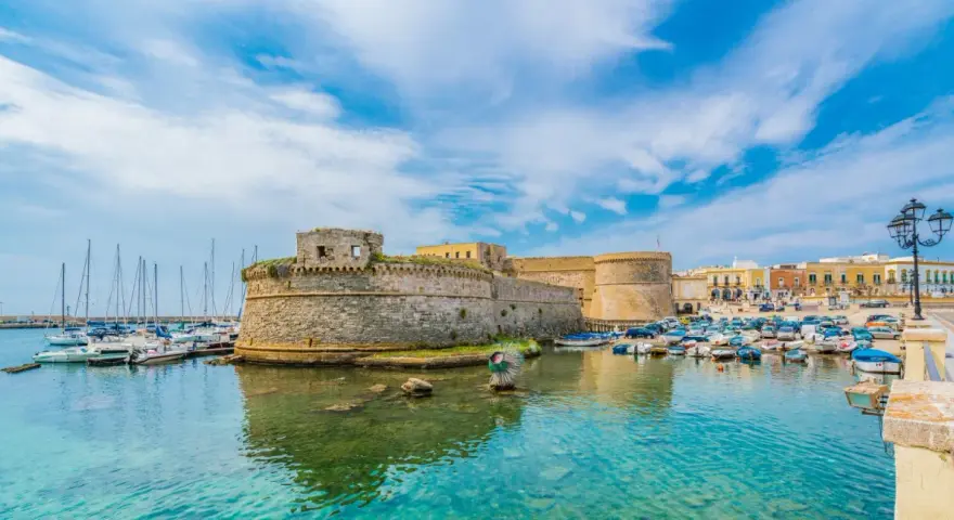 A stone castle overlooks a harbor filled with boats and yachts under a bright blue sky.