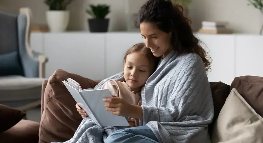 Mama czyta dziecku bajkę, otuleni kocem. Widać, ile jest opieki na dziecko w tej chwili.