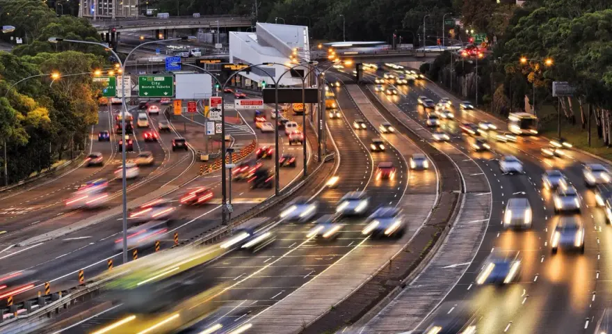 Traffico intenso su un'autostrada urbana al tramonto. Le luci dei veicoli creano scie luminose, con un **segnale 105** che indica la direzione.