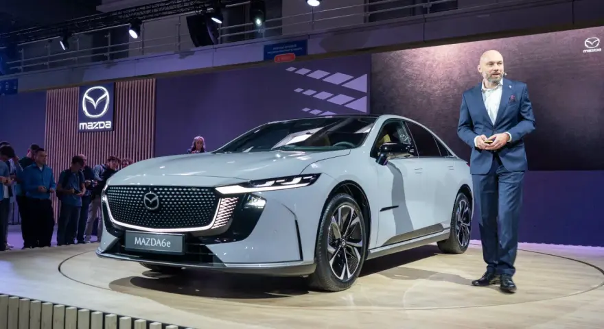 Elegancki, szary sedan Mazda6e prezentowany na scenie przez mężczyznę w garniturze.