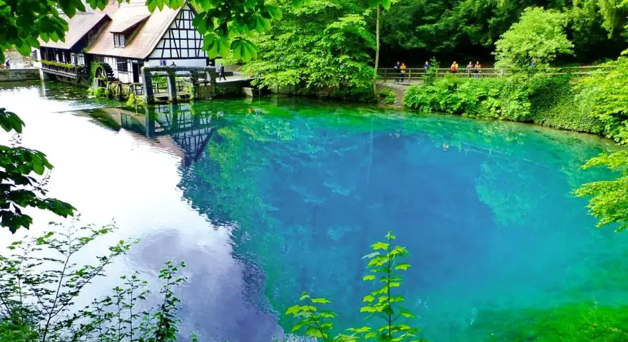 Der Blautopf See lädt zum Schwimmen ein. Ein historisches Fachwerkhaus mit Wasserrad spiegelt sich im kristallklaren, blauen Wasser.