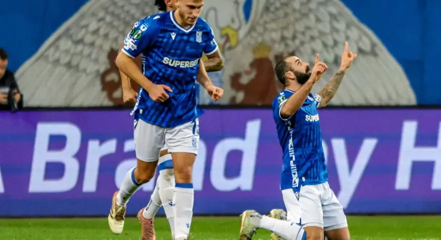 Piłkarz klęczy na boisku, celebrując bramkę. W tle biegnie kolega z drużyny. Wszyscy w niebieskich strojach. Czekamy, kiedy wraca ekstraklasa!