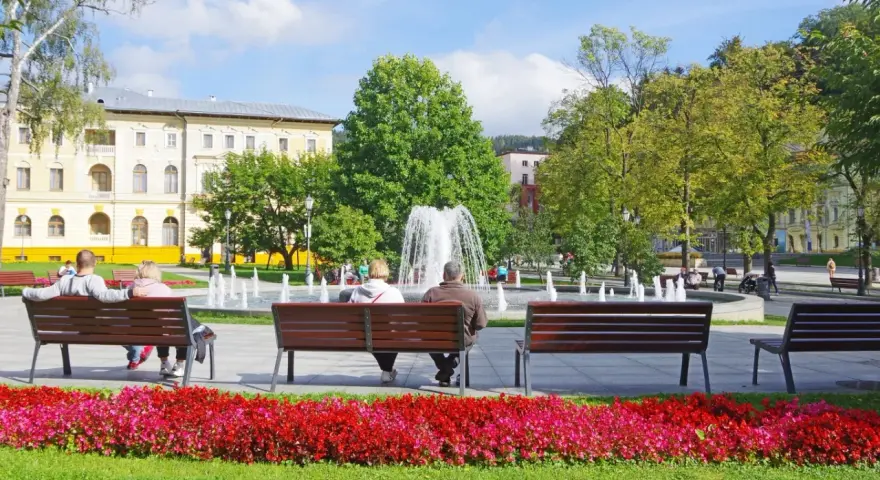 Park z fontanną i ludźmi siedzącymi na ławkach, w tle budynek.