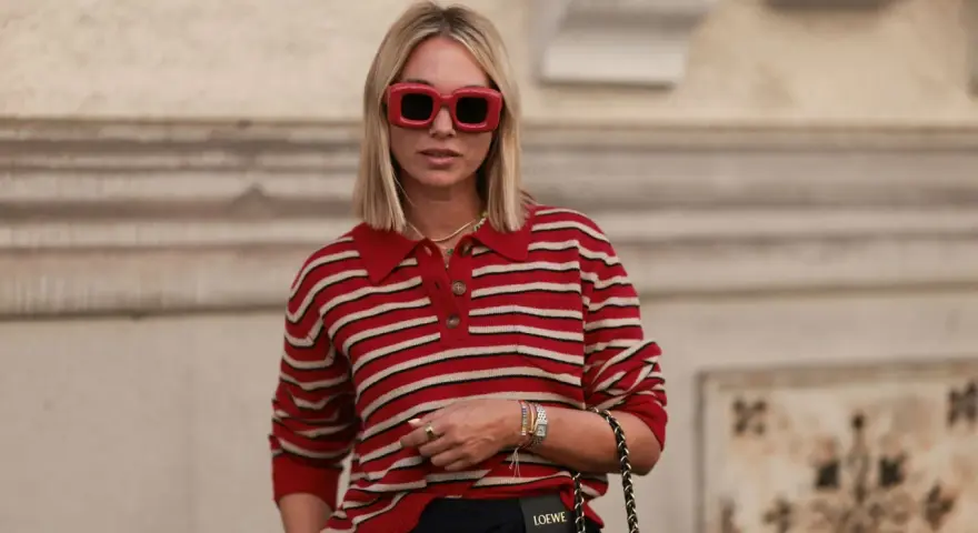 Donna con occhiali rossi oversize e maglione a righe rosse e bianche, un look che evoca un sofisticato preppy style.