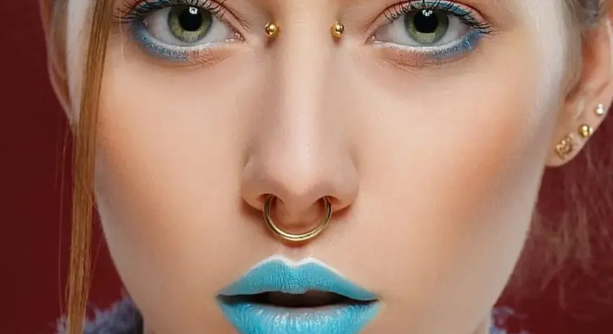 Kobieta z błękitnymi ustami i złotym septum. Jej oczy podkreślone są niebieskim cieniem, a między brwiami widoczny jest bridge piercing.