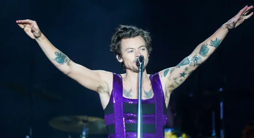 Harry Styles w błyszczącym fioletowym topie śpiewa na koncercie, unosząc ramiona. Jego ramiona pokryte są tatuażami.