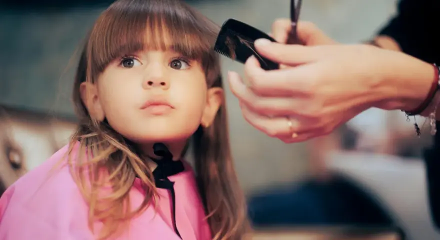Ein **Kind** bekommt einen neuen **Pony schneiden** beim Friseur. Die kleine Dame trägt einen pinken Umhang.