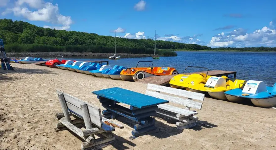Łódki wodne czekają na plaży nad jeziorem Sarbsko. Drewniane ławki i stół zapraszają do odpoczynku.