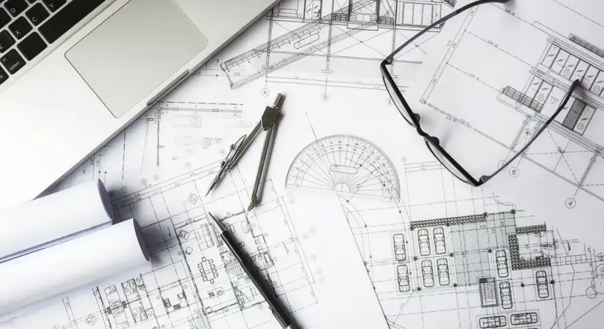 Na biurku architekta leżą plany budowlane, laptop, okulary i cyrkiel. Trwają prace nad przedmiarem robót.