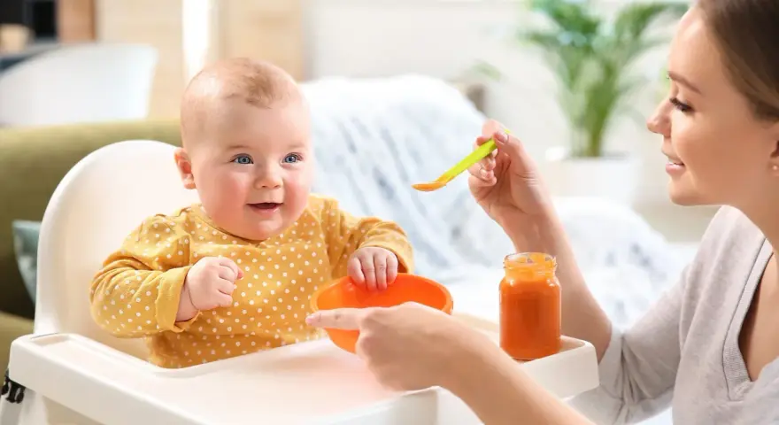 Uśmiechnięte 2 miesięczne dziecko w żółtym ubranku w kropki siedzi w krzesełku do karmienia. Mama podaje mu jedzenie z łyżeczki.