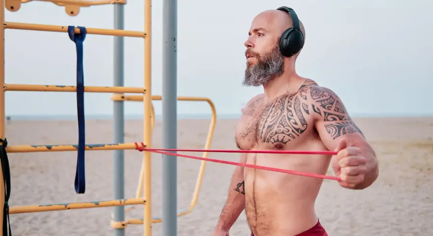Man med tatueringar och skägg tränar med gummisnoddar vid ett utegym på stranden.