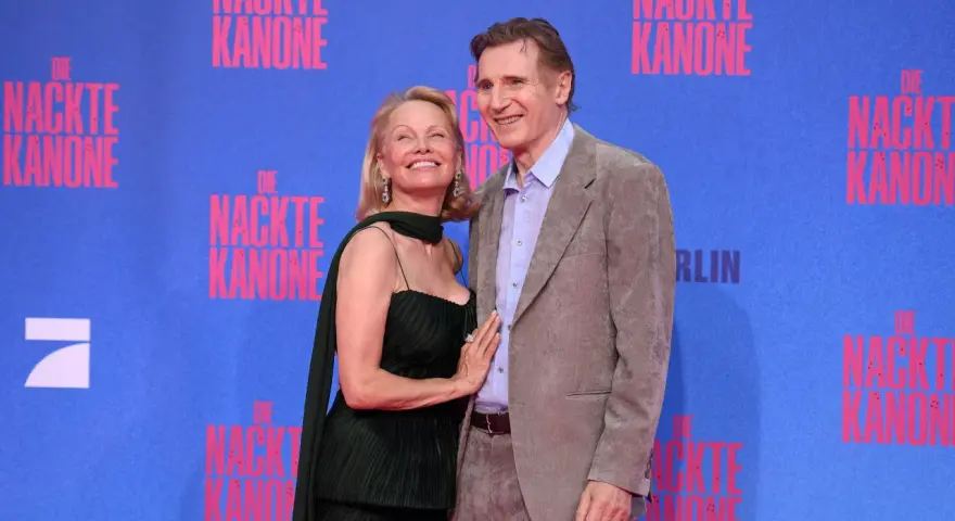 Liam Neeson e Pamela Anderson sorridono sul tappeto blu, con lo sfondo di "Die Nackte Kanone".