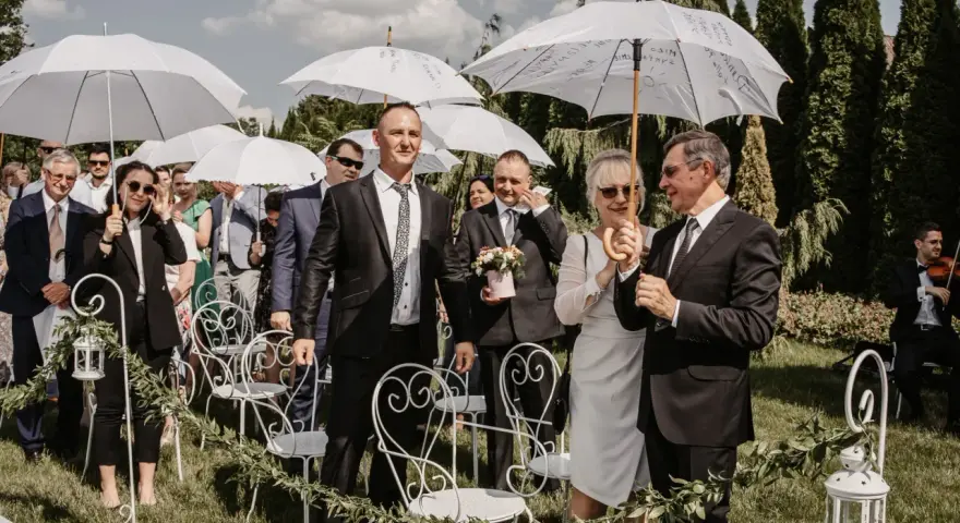 Wesele w kwietniu pod białymi parasolami. Goście w eleganckich strojach, wśród nich pan młody z bukiecikiem.