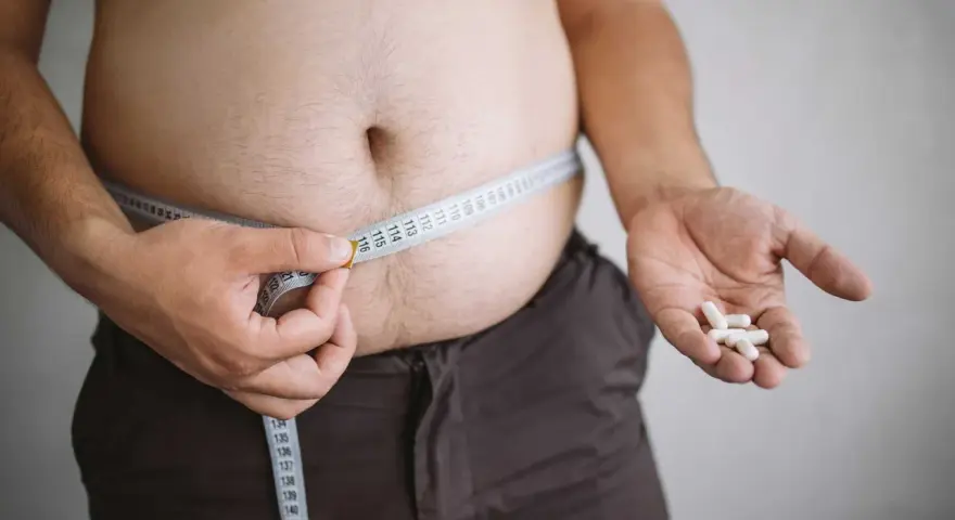 En person mäter sin midja med ett måttband, håller piller i handen. Kan indikera hormonell obalans och viktuppgång.