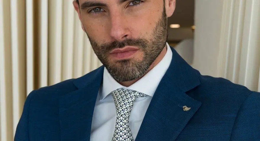 Uomo con abito blu, camicia bianca e cravatta con fantasia a pois bianchi e neri. Un tocco di stile con un pin a forma di ala.