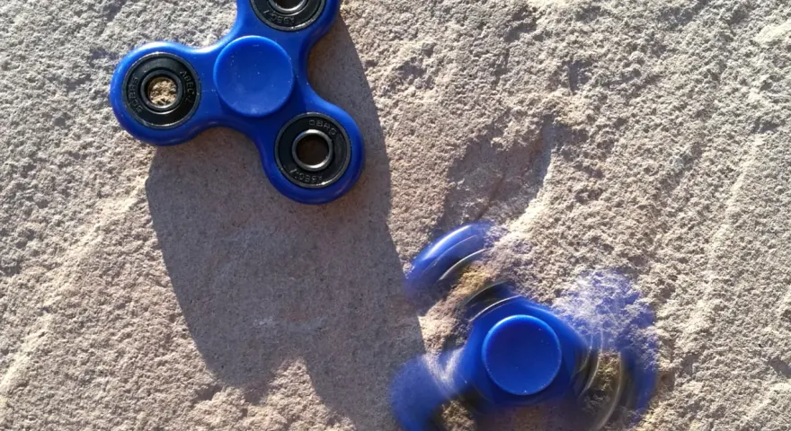 Dwa niebieskie fidget spinnery na piasku, jeden nieruchomy, drugi w ruchu, przypominają czasy, kiedy fidget spinnery były popularne.