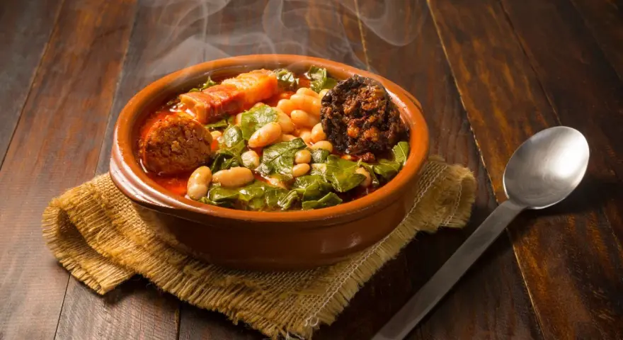 Un humeante pote asturiano, con fabes, berzas, chorizo y tocino, servido en cazuela de barro sobre arpillera.