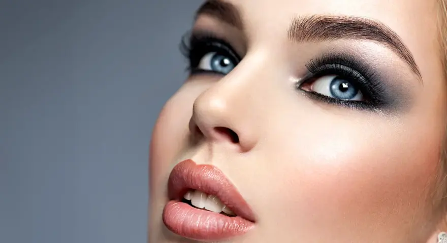 Niebieskie oczy z perfekcyjnym makijażem smokey eye, podkreślone długimi rzęsami i błyszczącymi ustami.