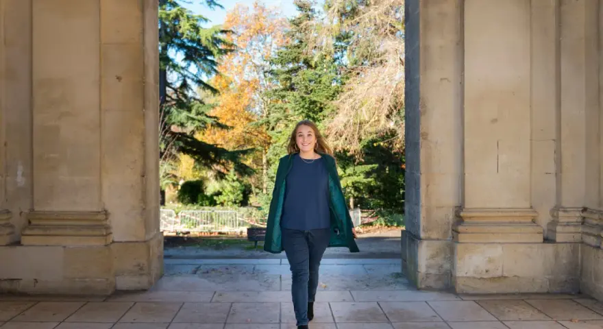 Une femme souriante marche sous une arche, portant un manteau vert et un haut bleu marine. L'automne colore les arbres en arrière-plan.