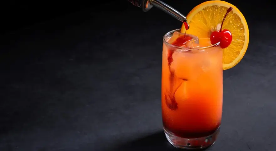 Kolorowy drink z syropem grenadina, ozdobiony plasterkiem pomarańczy i wisienką.