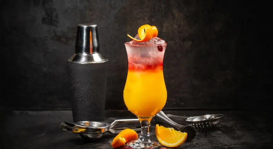 Kolorowy drink z lodem, pomarańczą i żurawiną, obok shakera. Wybór między Cosmopolitan a Sex on the Beach.