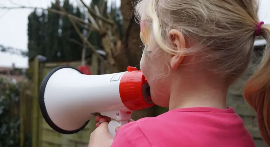 Ein Mädchen mit blonden Zöpfen hält ein Megafon und schreit hinein. Vielleicht fragt sie sich, warum sind Kinder so laut?