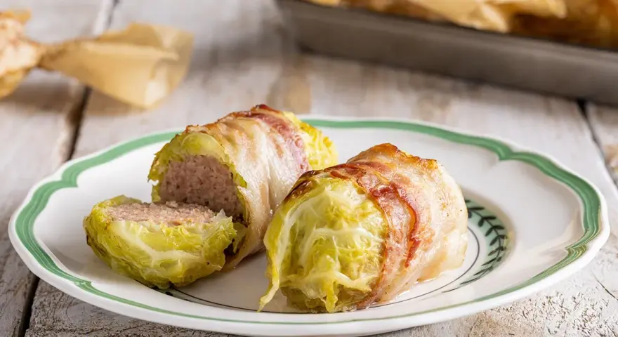 Deliziosi involtini di verza con carne, avvolti in pancetta croccante, serviti su un piatto bianco con bordo verde.