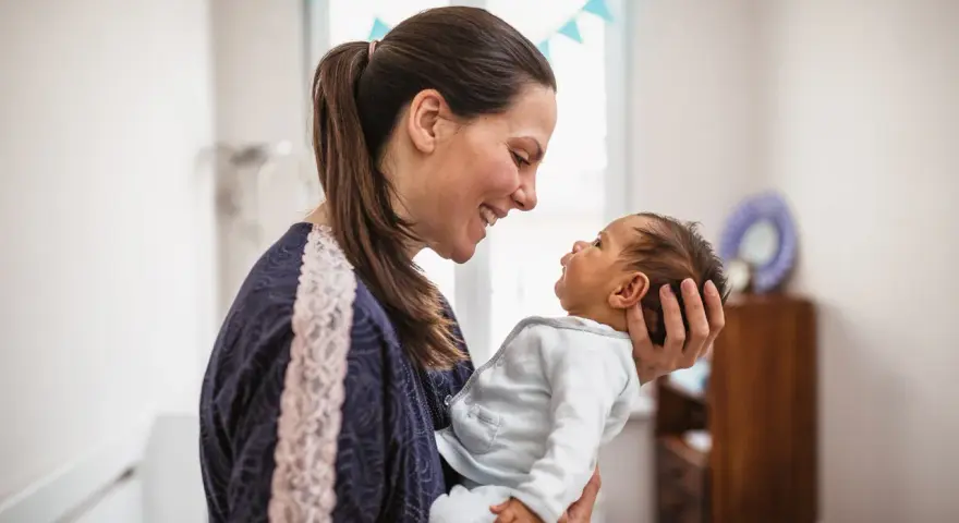 Uśmiechnięta mama trzyma na rękach noworodka. Dziecko patrzy na nią z zaciekawieniem, jakby miało zaraz kichnąć.