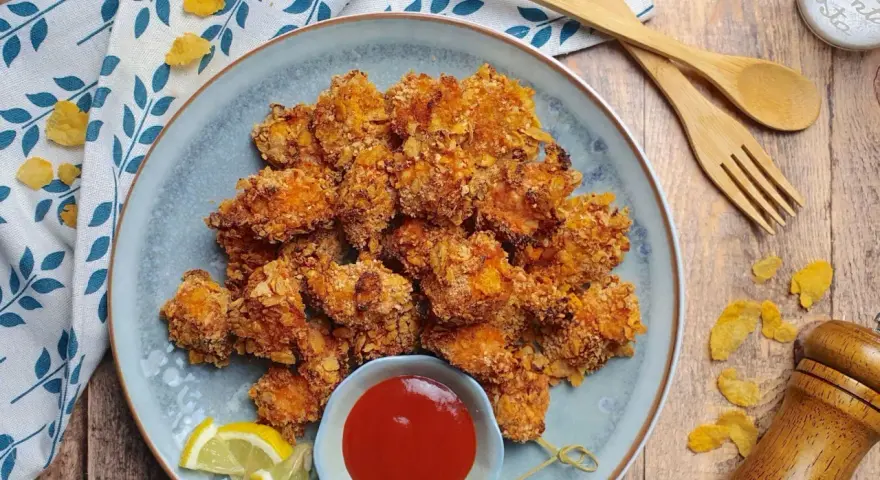 Piatto di croccanti bocconcini di pollo impanati con fiocchi di mais, serviti con ketchup e spicchi di limone.