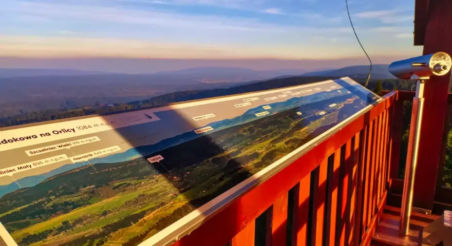 Widok z wieży widokowej na Orlicy. Na pierwszym planie panorama z opisem szczytów i dolin. W oddali łagodne wzgórza o zachodzie słońca.