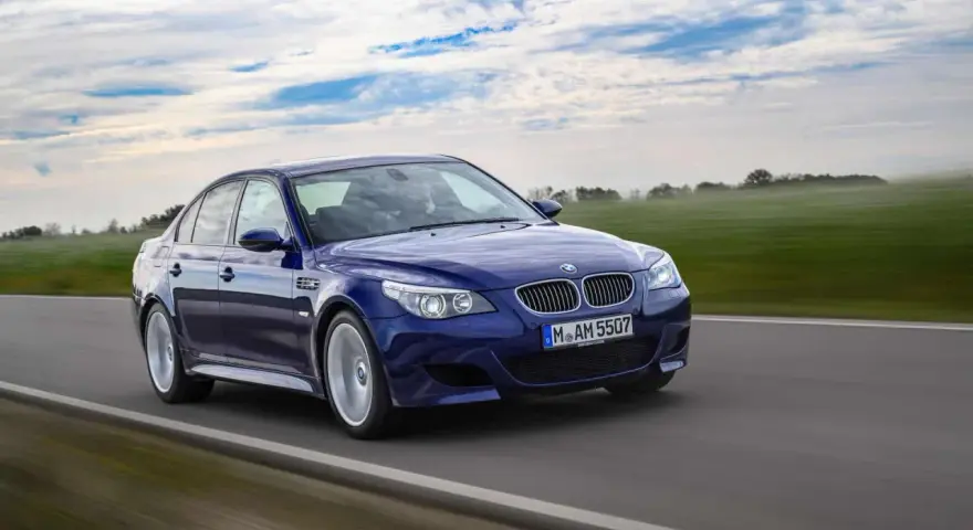 Niebieskie BMW M5 E60, ikona designu Chrisa Bangle'a, pędzi po drodze wśród zielonych pól i błękitnego nieba.