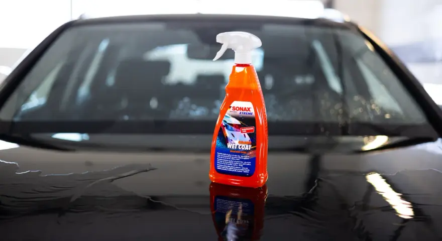 En flaska SONAX Xtreme Wet Coat står på en våt, mörk bilhuv.