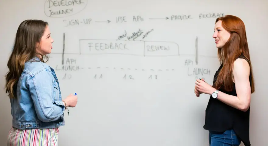 Dos mujeres discuten un diagrama de "Developer Journey" en una pizarra. Una profesora explica el proceso.